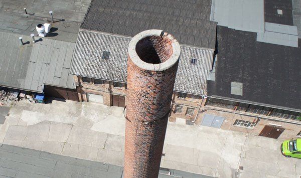 Drohnenaufnahme eines alten Industrieschornsteins aus Backstein mit Rissen, Blick auf das Dach einer Fabrikhalle.