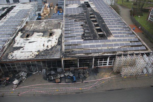 Luftaufnahme eines Industriegebäudes mit verbranntem Flachdach und beschädigten Solarpanelen nach einem Brand.