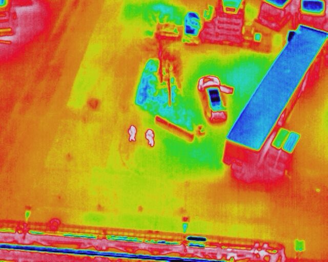 Thermal image of an area with cars, storage spaces, and two visible people.