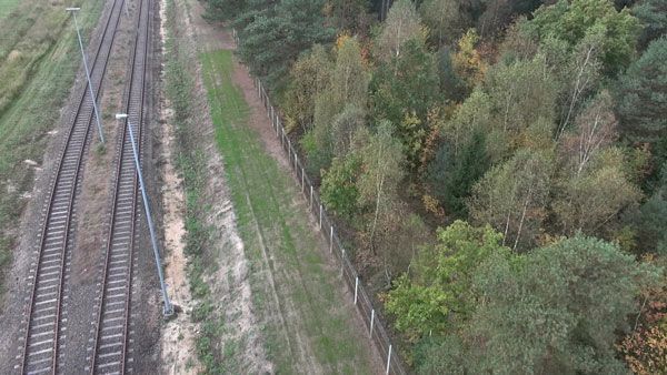 Aerial view of railway tracks next to forest and security fence, showing track bed and vegetation contrast.