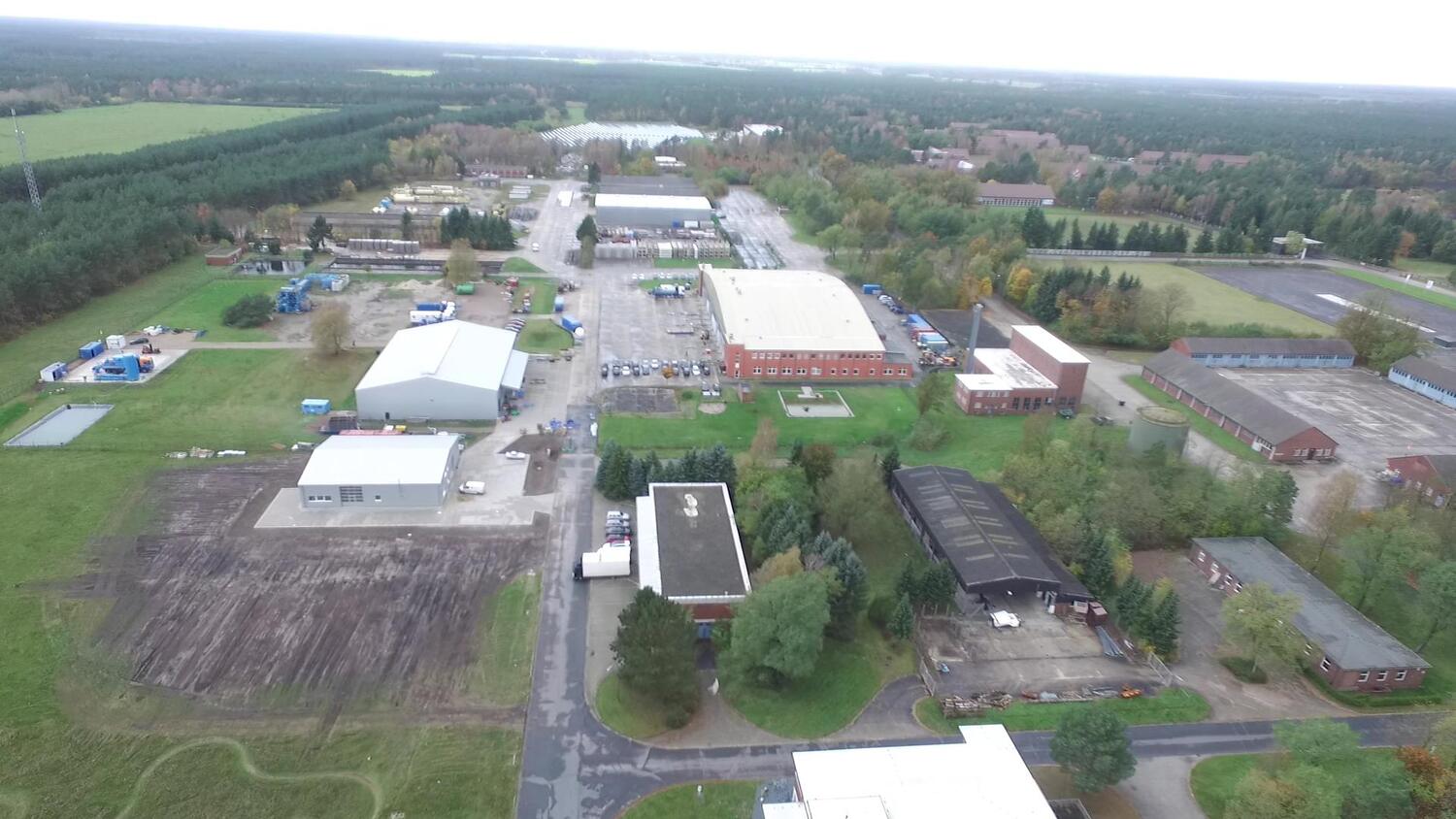 Drohnenaufnahme eines weitläufigen Industrie- und Gewerbeareals mit Hallen, Parkplätzen, Straßen und angrenzenden Waldflächen.