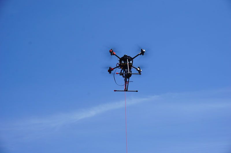 Multikopter-Drohne mit Kabelverbindung schwebt vor blauem Himmel.